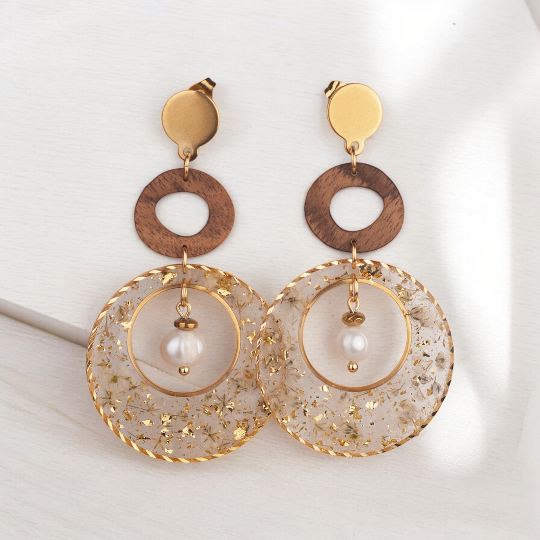 Handgefertigte Ohrringe aus Nussholz mit rundem Resin-Element mit getrockneten weißen Blüten und Blattgold, Süßwasserzuchtperle und vergoldetem Edelstahl-Ohrstecker auf hellem Hintergrund.