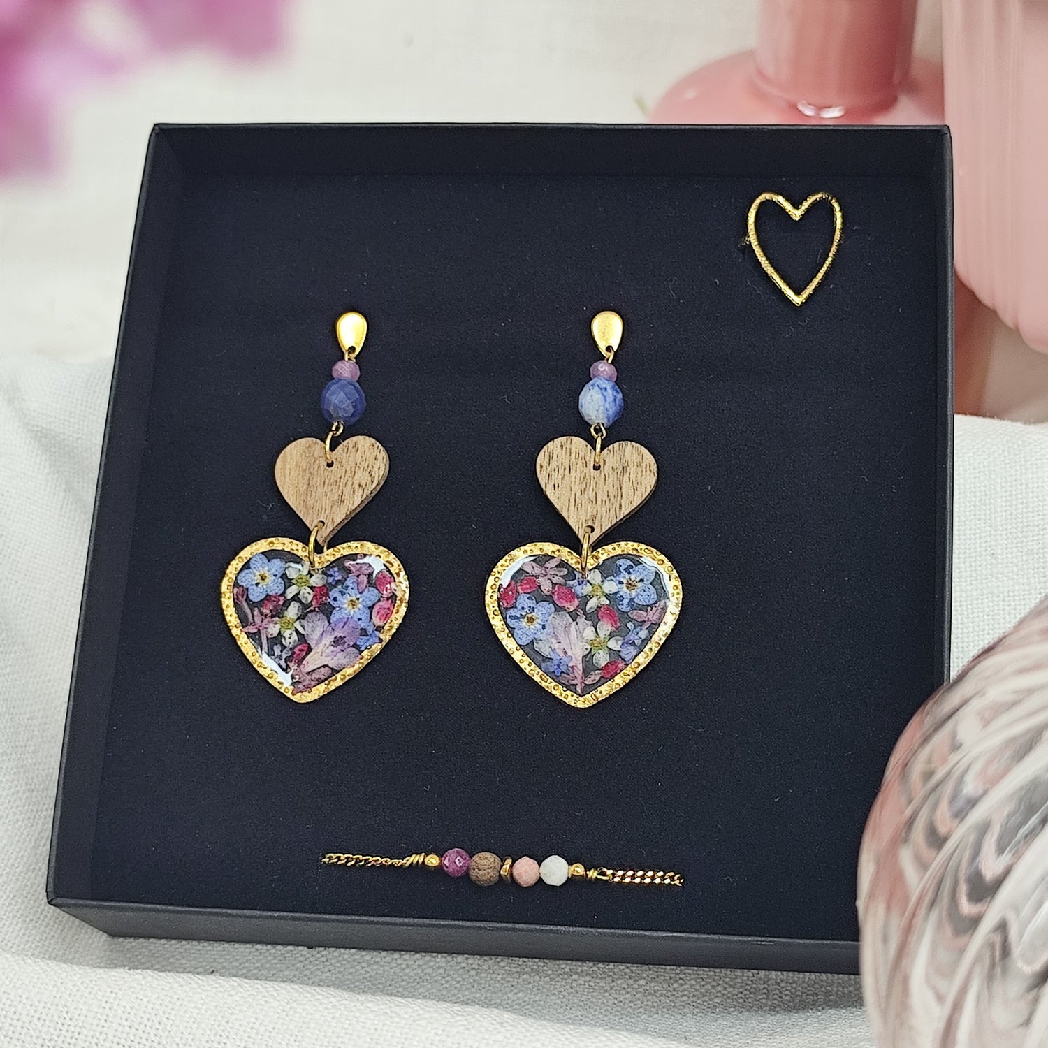 Schmuckset mit herzförmigen Ohrringen mit Resin-Elementen mit echten Blüten, Edelstahlring gold in Herzform und feinem Armketterl mit Natursteinen, arrangiert in einer schwarzen Schmuckschachtel. Rosa Vasen im Vordergund
