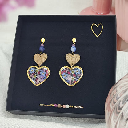 Schmuckset mit herzförmigen Ohrringen mit Resin-Elementen mit echten Blüten, Edelstahlring gold in Herzform und feinem Armketterl mit Natursteinen, arrangiert in einer schwarzen Schmuckschachtel. Rosa Vasen im Vordergund