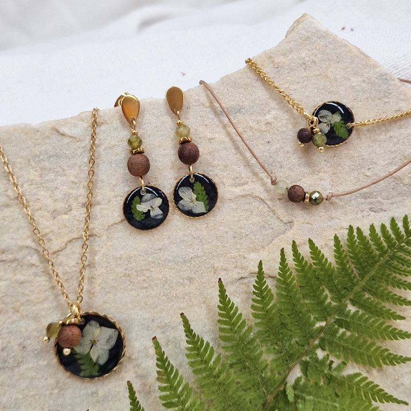 Handgefertigtes Schmuckset ‚Mini Waldlicht‘ aus Ohrringen, Halskette, Armkettchen und Armband mit schwarzem Resin-Element mit echten getrockneten Blüten und Farnen, Nussholz, Natursteinen und goldfarbenen Edelstahldetails, arrangiert auf Naturstein