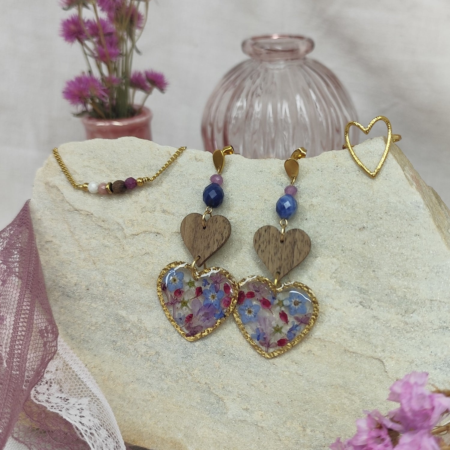 Schmuckset mit herzförmigen Ohrringen mit Resin-Elementen mit echten Blüten, Edelstahlring gold in Herzform und feinem Armketterl mit Natursteinen, arrangiert auf hellem Stein nebeneinander. Rosa Vasen und Blumen im Vordergund