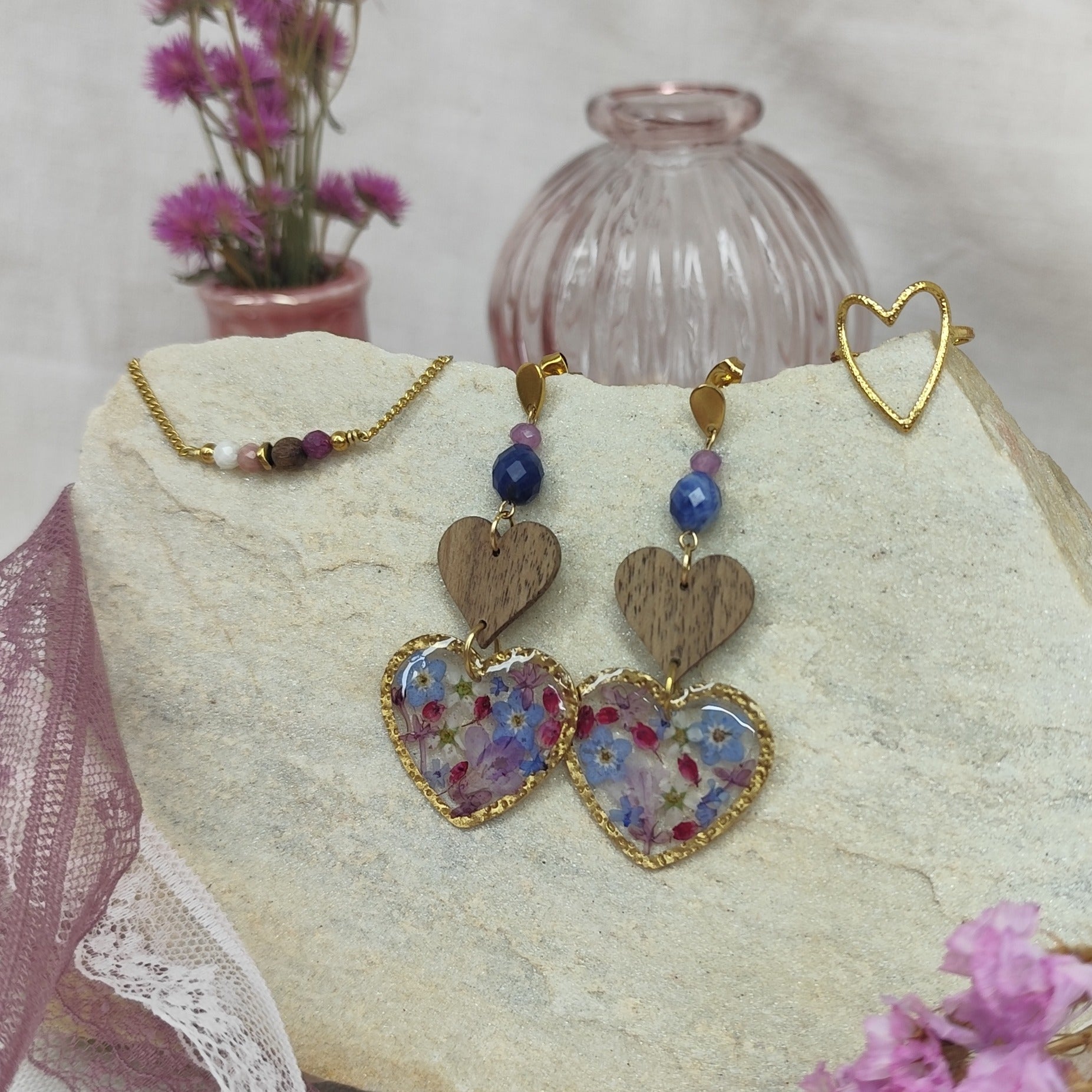 Schmuckset mit herzförmigen Ohrringen mit Resin-Elementen mit echten Blüten, Edelstahlring gold in Herzform und feinem Armketterl mit Natursteinen, arrangiert auf hellem Stein nebeneinander. Rosa Vasen und Blumen im Vordergund