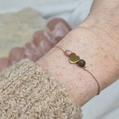 Armband mit selbstgemachter Holzperle, Rhodochronit Stein und goldenem Herz auf Handgelenk getragen. Rosa Hintergrund