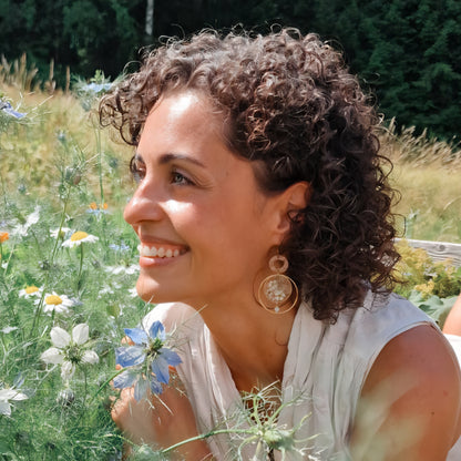 Frau mit braunen, schulterlangen Locken liegt auf sommerlicher Blumenwiese trägt handgefertigte Holzohrringe mit echten gepressten Blüten im transparenten Resin, Nussholz-Element und kleiner Perle.