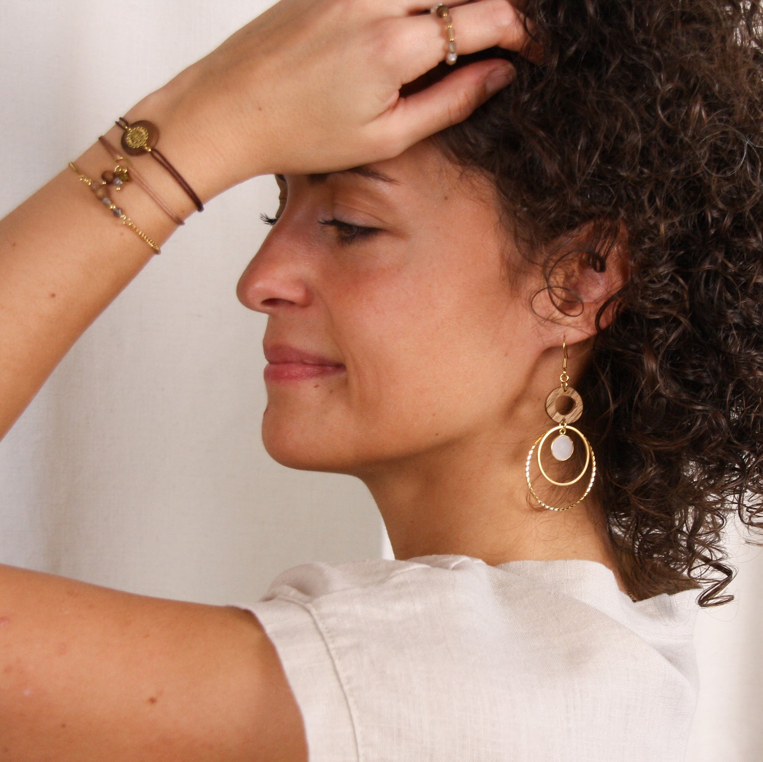 Frau trägt die Ohrringe ‚Harmonia‘ aus Nussholz, weißem Quarz und goldfarbenen Metallelementen sowie eine kombinierte Harmonia-Armbandkombination am Handgelenk