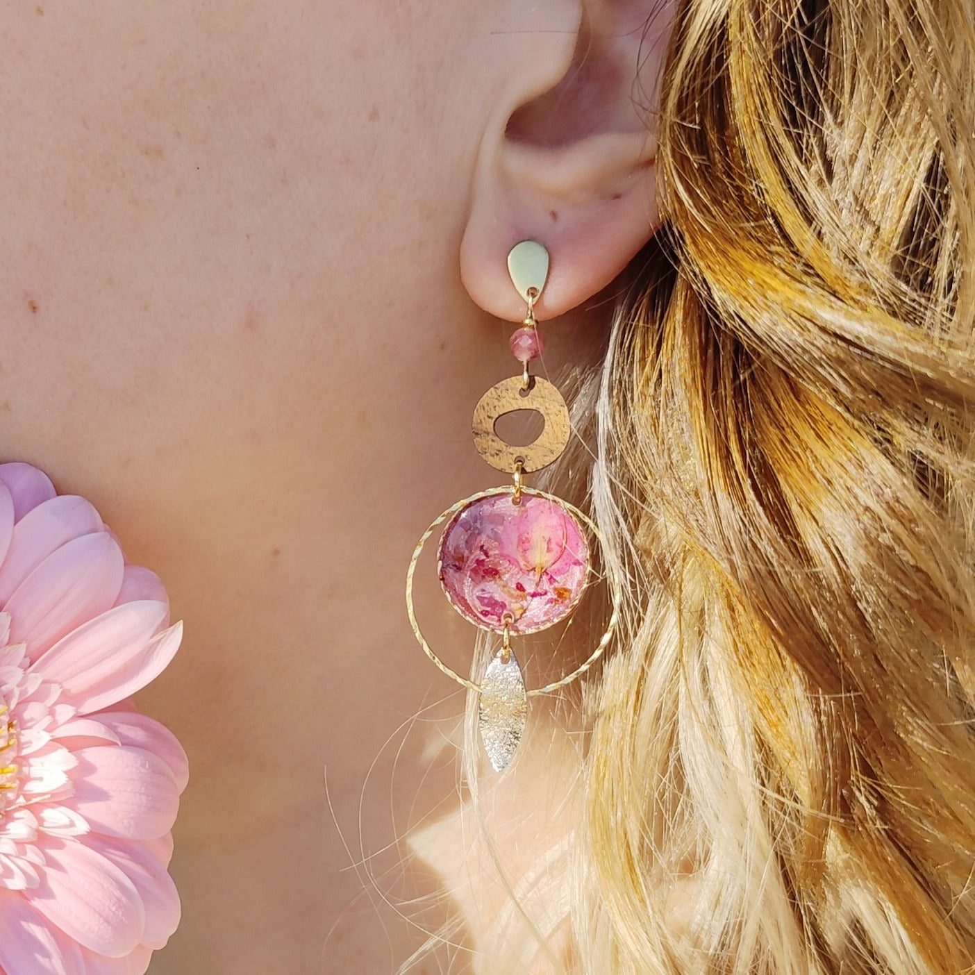 Am Ohr getragener Ohrring aus goldfarbenem Edelstahl mit Nussholzscheibe, rundem Resin-Element mit echten rosa Blüten und feinem Metallelement, seitlich im Sonnenlicht