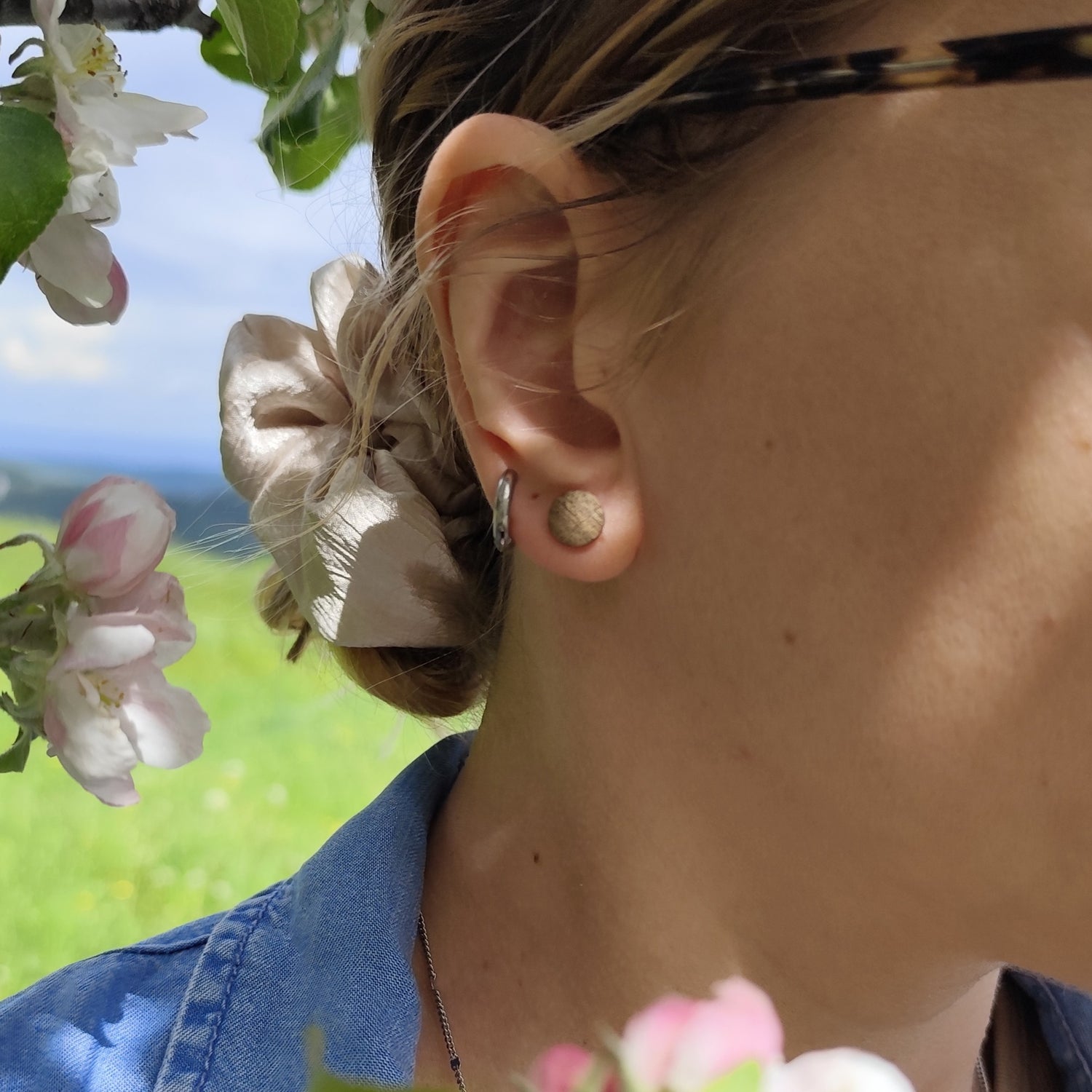 Person trägt einen runden Ohrstecker aus braunem Holz am Ohr. Es scheint die Sonne und links oben sind Kirschblüten