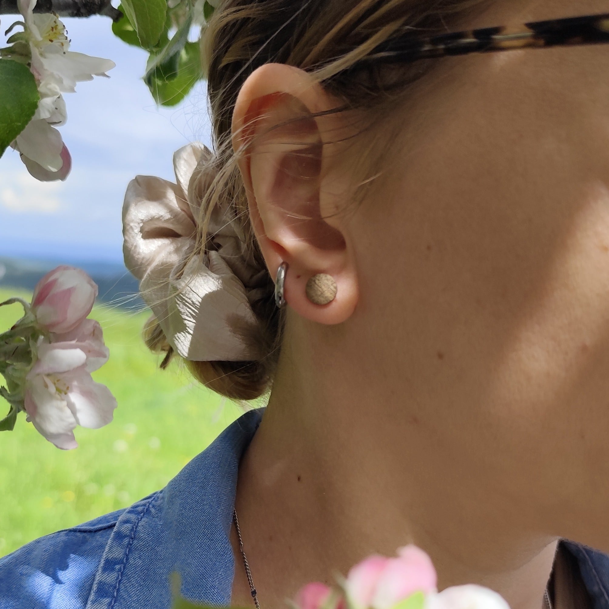 Person trägt einen runden Ohrstecker aus braunem Holz am Ohr. Es scheint die Sonne und links oben sind Kirschblüten