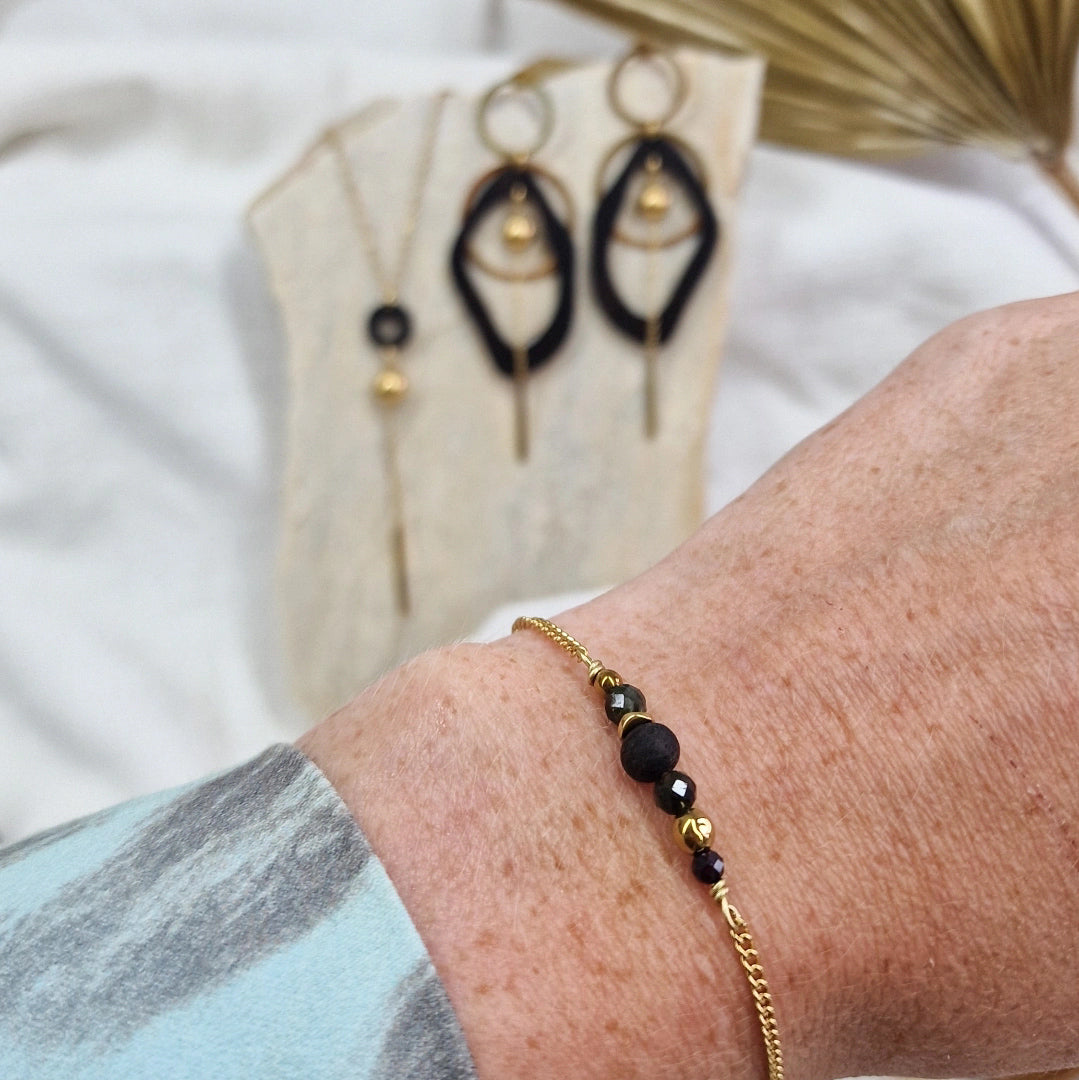 Handgefertigtes Armband aus dunklen Holzperlen mit goldfarbener Kette am Handgelenk getragen, im Hintergrund passende Ohrringe und Halskette