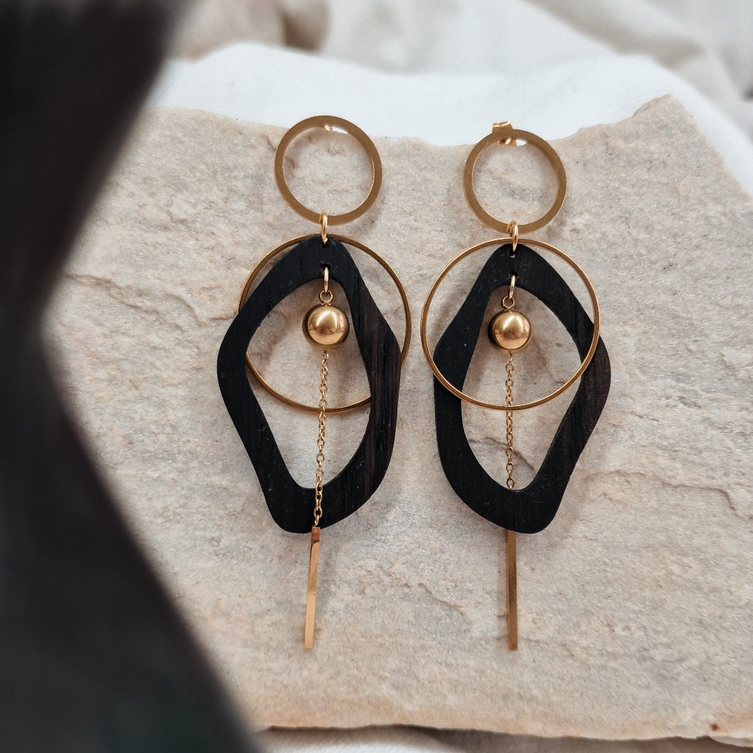 Paar Ohrringe aus dunkler Räuchereiche mit ovaler Holzform, kreisförmigem Metallelement und feiner Kette, liegend auf hellem Stein
