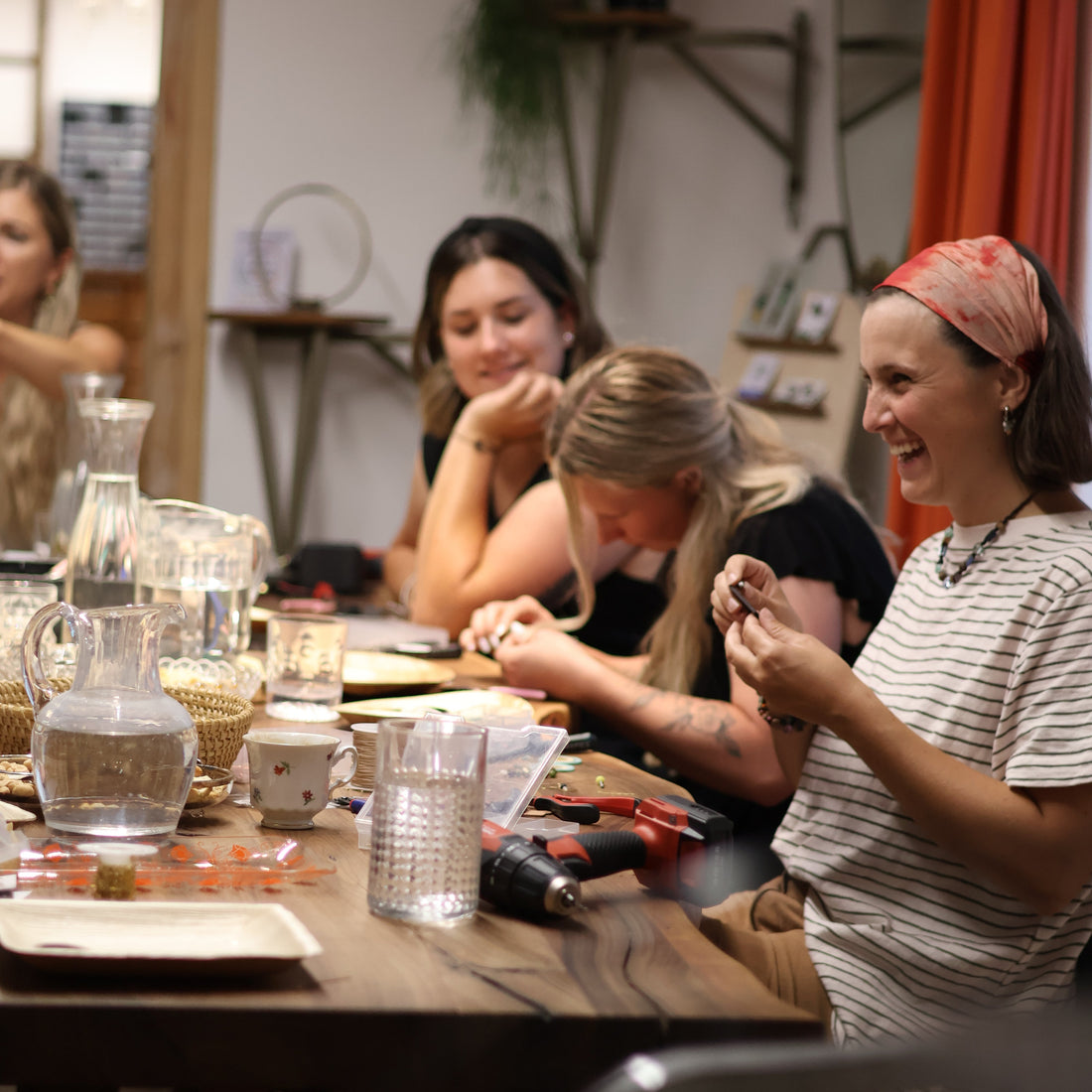 Lachende Person im Vordergrund, 3 Personen im Hintergrund arbeiten konzentriert an ihren Schmuckstücken. Ausgelassene Stimmung während eines Workshops, alle sitzen am Tisch